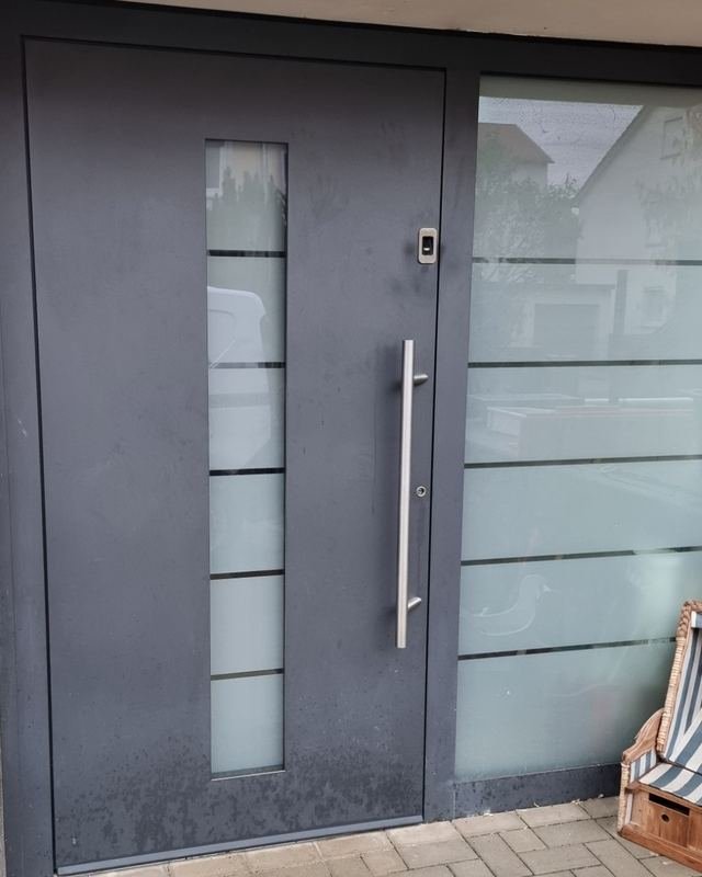 Anthrazitfarbene Haustür mit langem Edelstahlgriff und schmalem Glasstreifen, daneben eine Milchglasscheibe
