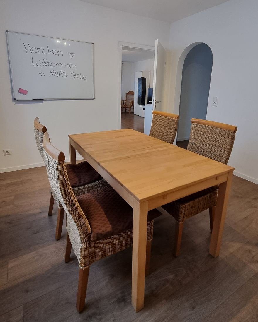 Esszimmer mit Holztisch, vier Korbstühlen und einem Whiteboard mit der Aufschrift „Herzlich Willkommen in Arinas Stüble“, durch die offene Tür sieht man ins Wohnzimmer