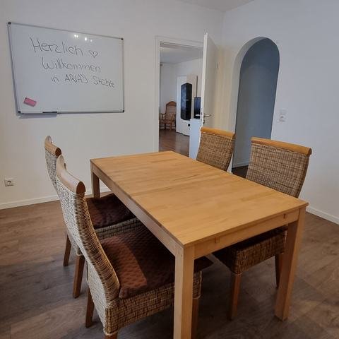 Esszimmer mit Holztisch und Whiteboard „Herzlich Willkommen in Arinas Stüble“, dahinter die offene Tür zum Wohnzimmer