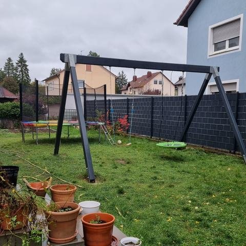 Garten mit Rasen, Schaukelgestell mit Nestschaukel, Trampolin im Hintergrund und Blumentöpfen