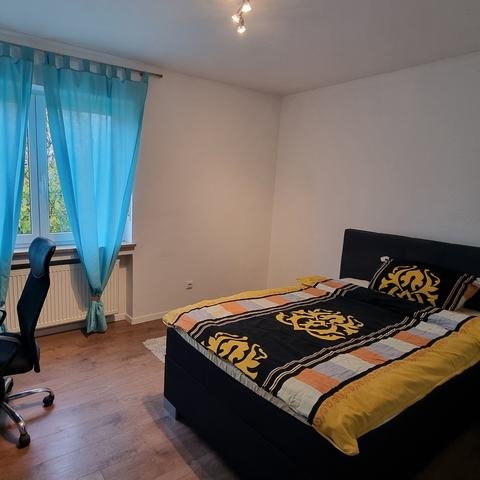 Schlafzimmer 1 mit bezogenem dunklem Boxspringbett, Nachttisch, weißem Schreibtisch mit Bürostuhl und türkisen Vorhängen