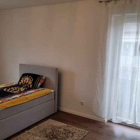 Schlafzimmer 2 mit hellgrauem Boxspringbett und einer hohen Glastür mit weißen Vorhängen