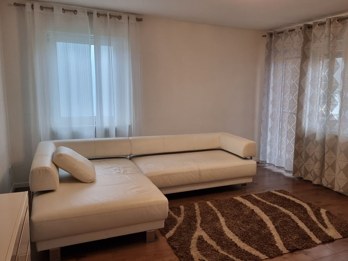 Helles Wohnzimmer mit weißem Ecksofa aus Leder, braun gemustertem Teppich und hellen Vorhängen vor den Fenstern
