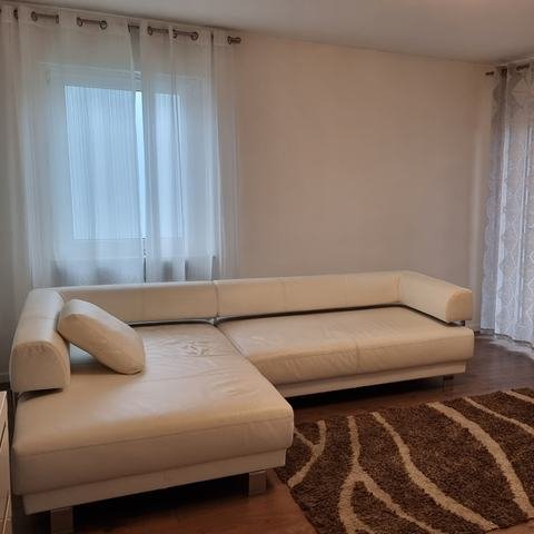 Wohnzimmer mit weißem Ecksofa aus Leder, braun gemustertem Teppich und hellen Vorhängen